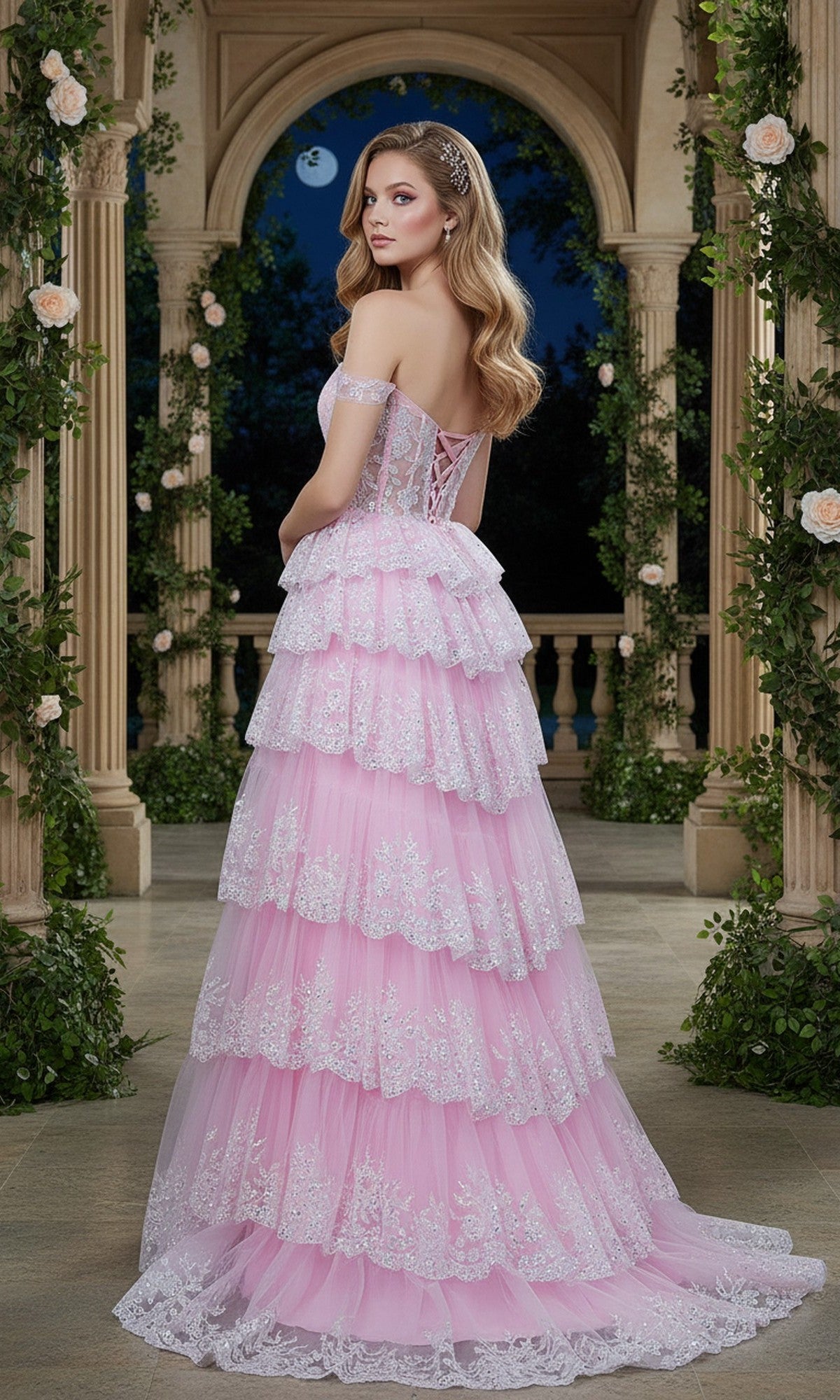 Wearing the Cinderella Couture 8499J, a woman in a tiered, off-the-shoulder light pink prom dress with a corset bodice and lace accents stands in an ornate garden pavilion at night, turned slightly from the camera.